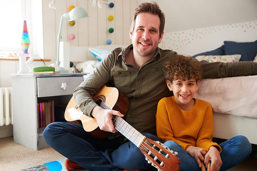 Personal Insurance - Father and Son Sit on Son's Bedroom Floor, Dad Holding an Acoustic Guitar, Both Smiling